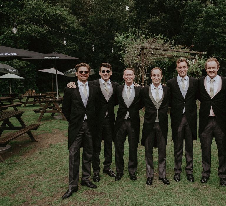 Groom and groomsmen stand arm in arm in black morning coats, coloured waistcoats and bespoke olive leaf print ties before Surrey summer wedding