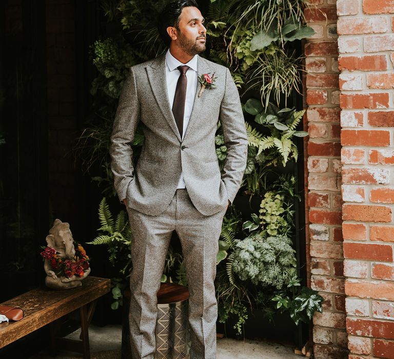 An Indian groom wearing a grey suit stands outside his wedding venue. 