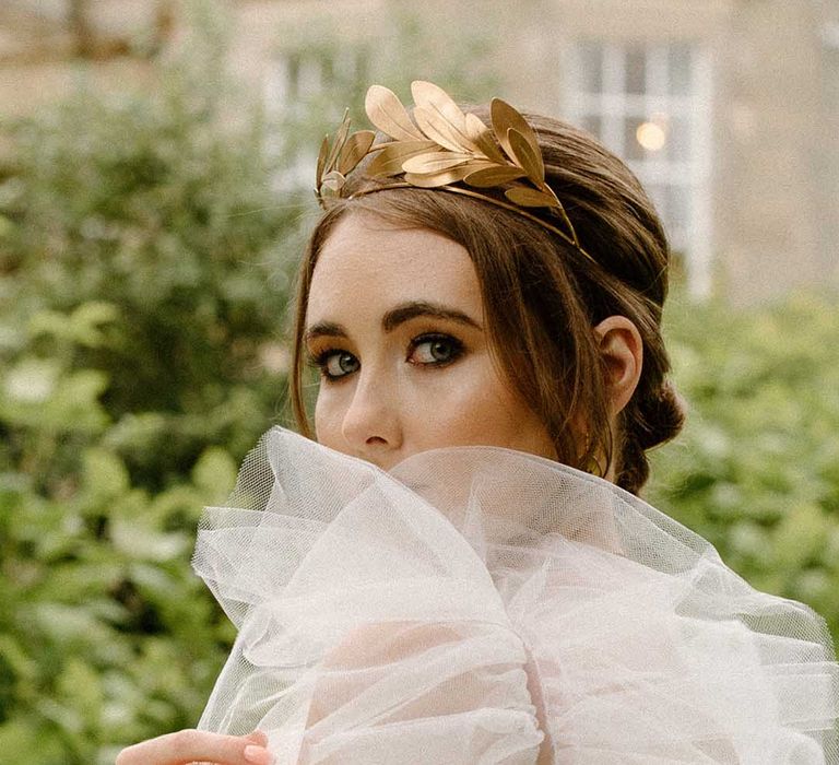 Beautiful bride with black eyeliner looking through her tulle puff sleeves wearing a gold headdress 