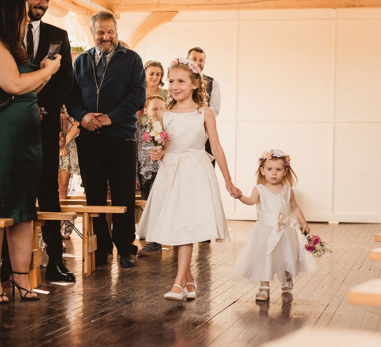 Two flower girls in white dresses and flowers crowns holding small wedding bouquets walk down the aisle at Inkersall Grange Farm wedding ceremony