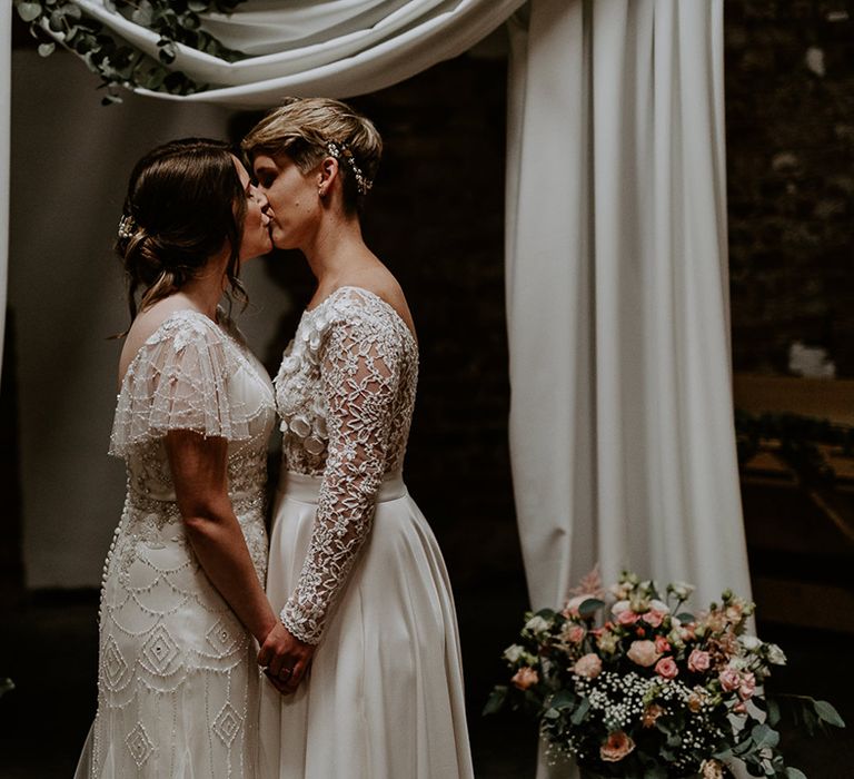 Portrait of two brides in a long sleeve wedding dress and vintage inspired gown kissing at their draped altar at Woolas Barn