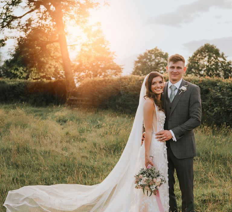 Bride in applique Berta wedding dress with train and veil holding mixed bridal bouquet stands in field hugging groom in grey checked three piece suit during golden hour for summer wedding at Primrose Hill Farm