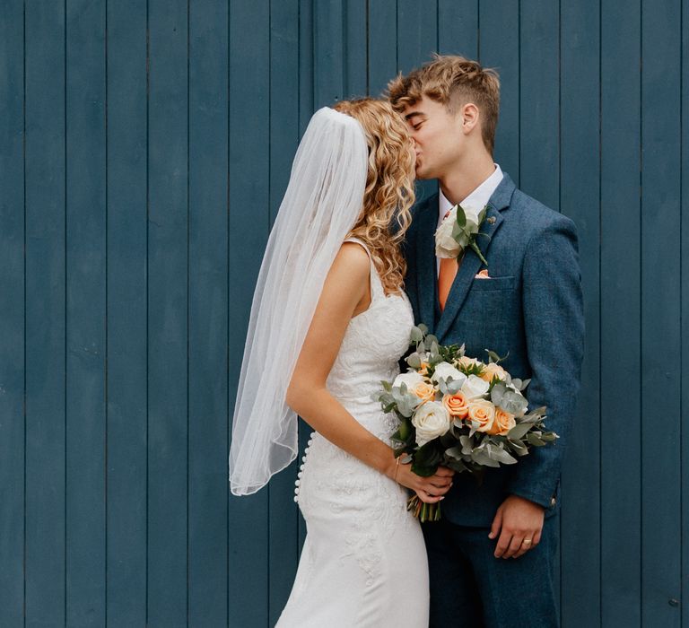 Bride & groom kiss after wedding ceremony in front of blue door whilst bride holds floral bouquet complete with peach flowers 