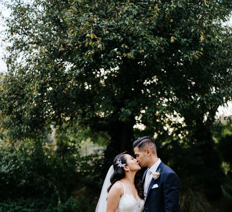 Groom in a navy suit kissing his bride in an sequin wedding dress and cathedral length veil as she holds a pink wedding bouquet 