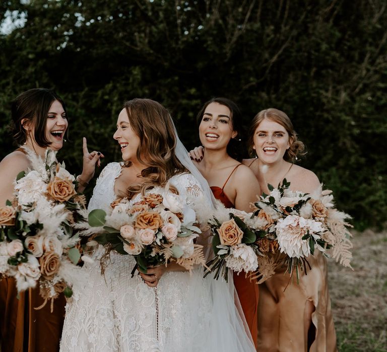 Bride in homemade lace wedding dress and veil stands with bridesmaids in assorted silk bridesmaid dresses as they all hold mixed rose wedding bouquets for outdoor wedding in Bedfordshire