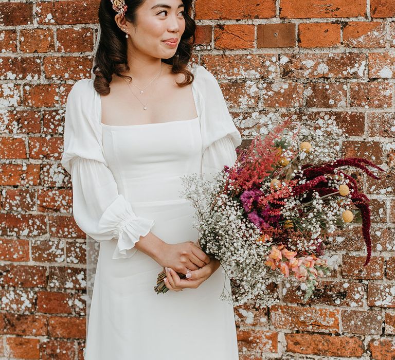 Bride stands in front of brick wall as she holds colourful homemade floral bouquet