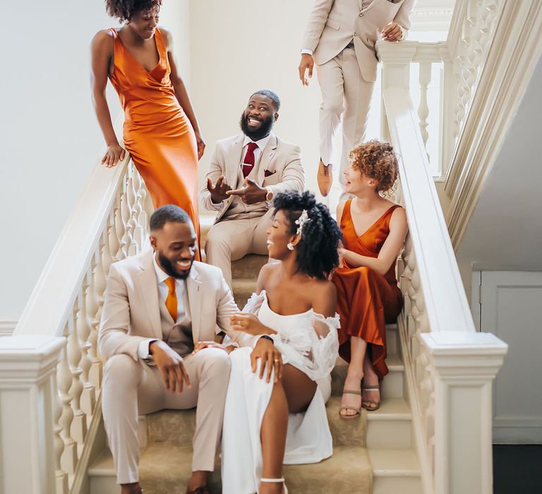 Wedding party in beige and orange suits and satin bridesmaid dresses sitting no the stairs at Modern Hall, London