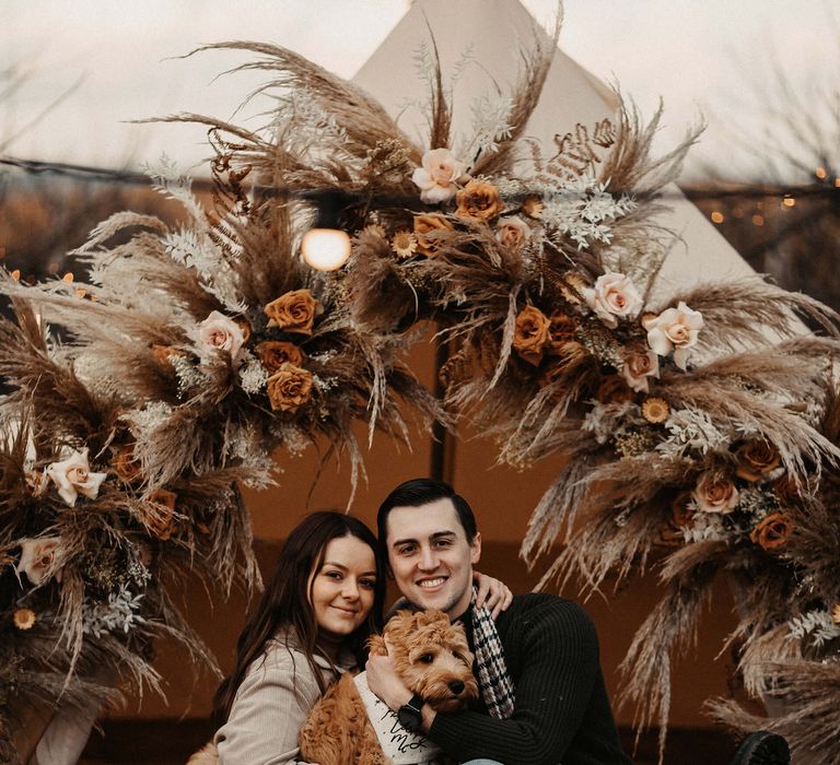 Bride and groom to be sitting in front of a bell tent decorated with dried flower with their pet cockapoo 