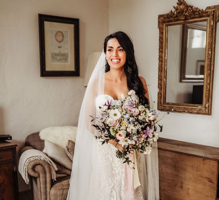 Bride in lace and tulle wedding dress with sweetheart neckline and cathedral veil stands holding pastel summer bridal bouquet