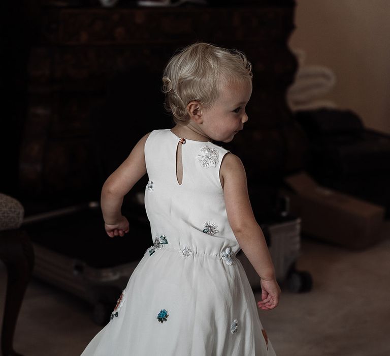 Flower girl in white dress with floral embroidery stands in bedroom before wedding