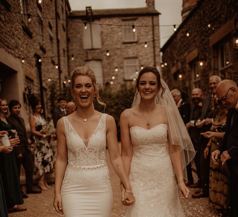 Brides walk hand in hand on their wedding day as guests watch 