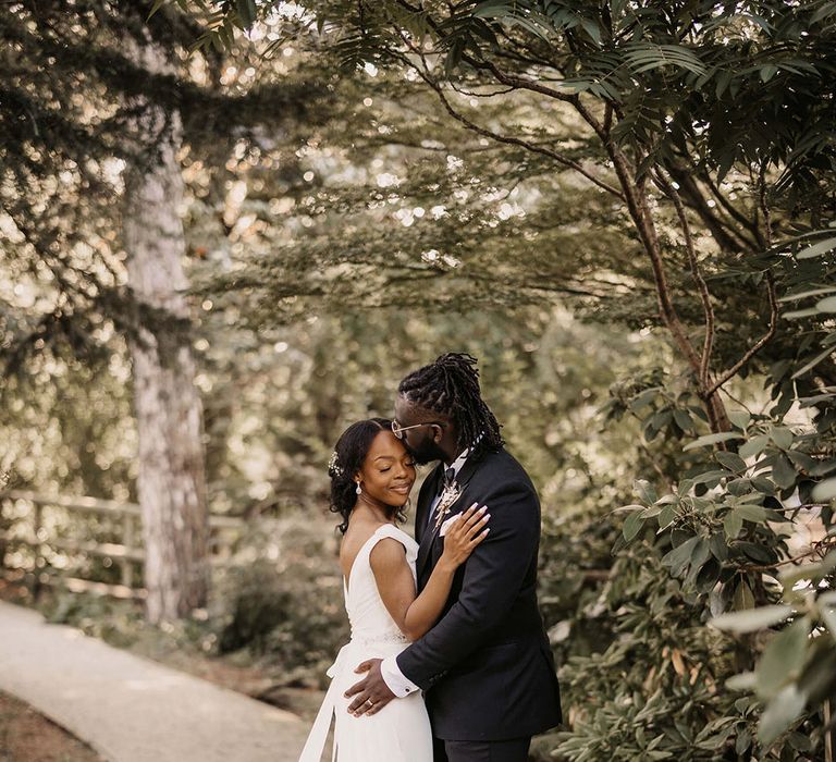 Bride & groom stand outdoors under trees on their wedding day