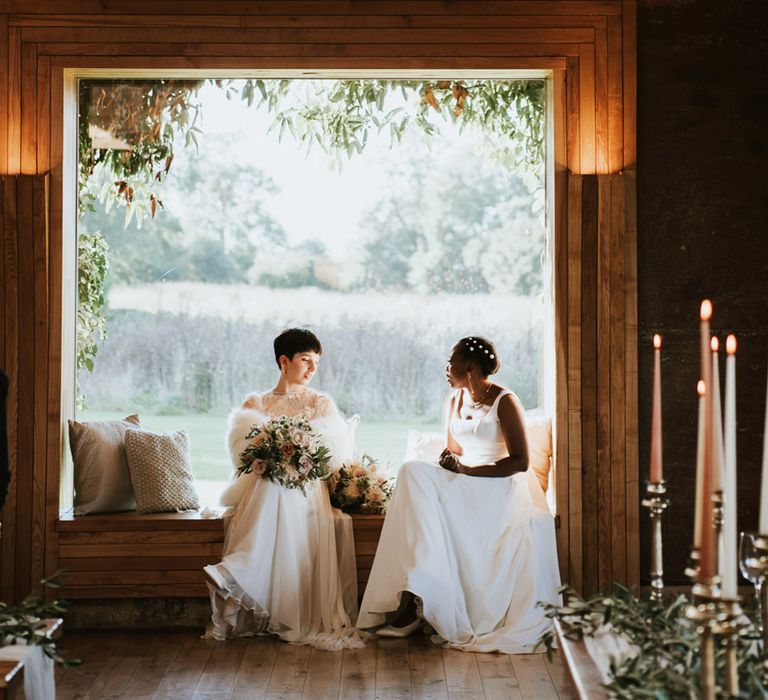 Bride in a Anna Kara wedding dress and faux fur stole sitting on a bench with her Black bride in a Essense of Australia wedding dress 