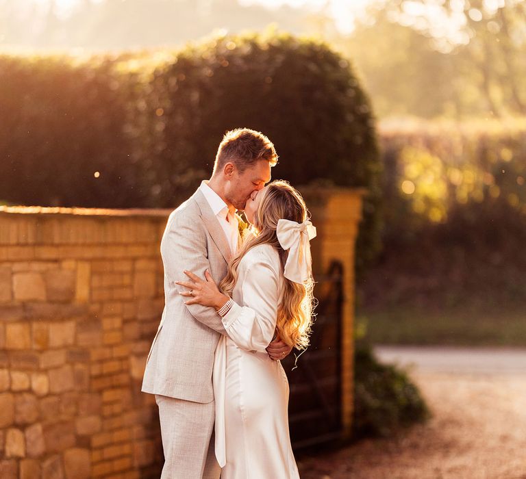 Bride with satin hair bow and satin wedding dress with train stands kissing groom in light grey suit and brown suede shoes during golden hour