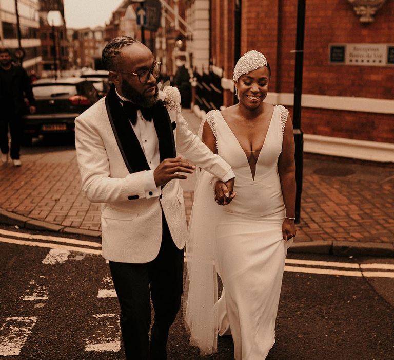 Groom in a white brocade tuxedo jacket leading his bride through the streets of Birmingham in a fitted wedding Dres with lunging neckline and a beaded cap vintage style headdress 