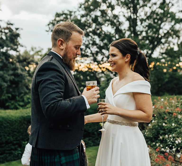 Bride in an off the shoulder Suzanne Neville wedding dress and groom in a tartan kilt drinking cocktail at sunset 