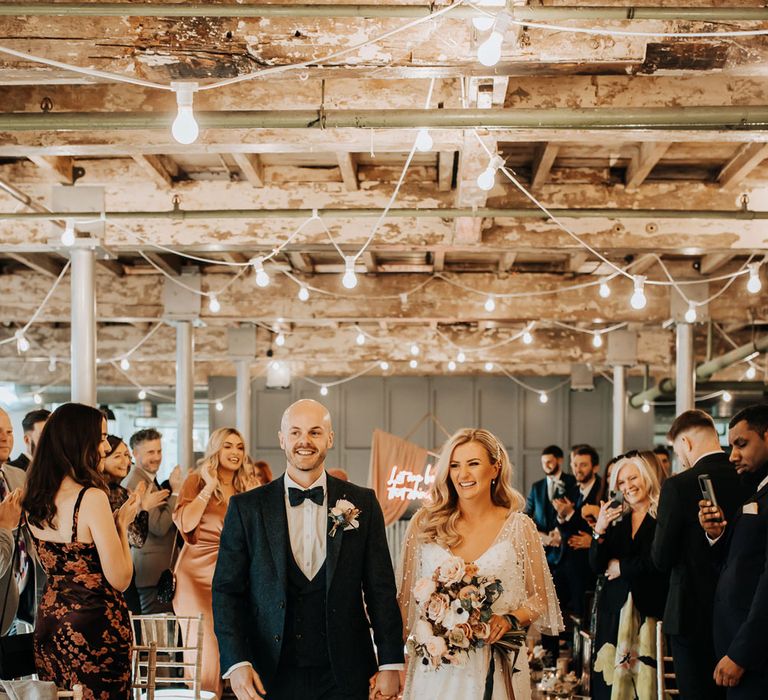 Bride in pearl wedding dress holding pastel bouquet tied with ribbon walks back down aisle with groom in three piece navy wedding suit with bow tie at industrial Mill wedding