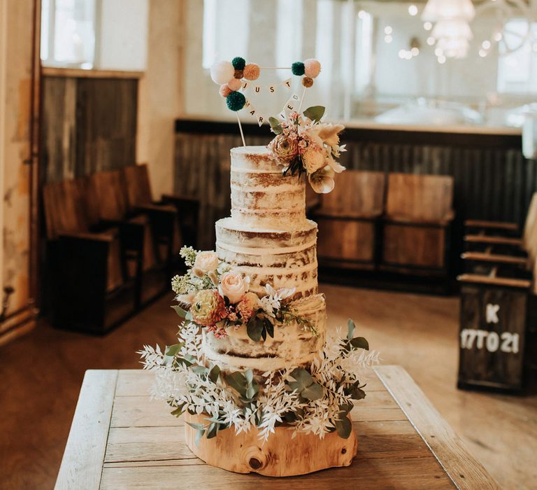 Rustic semi naked three tier wedding cake with floral decor and bunting wedding topper