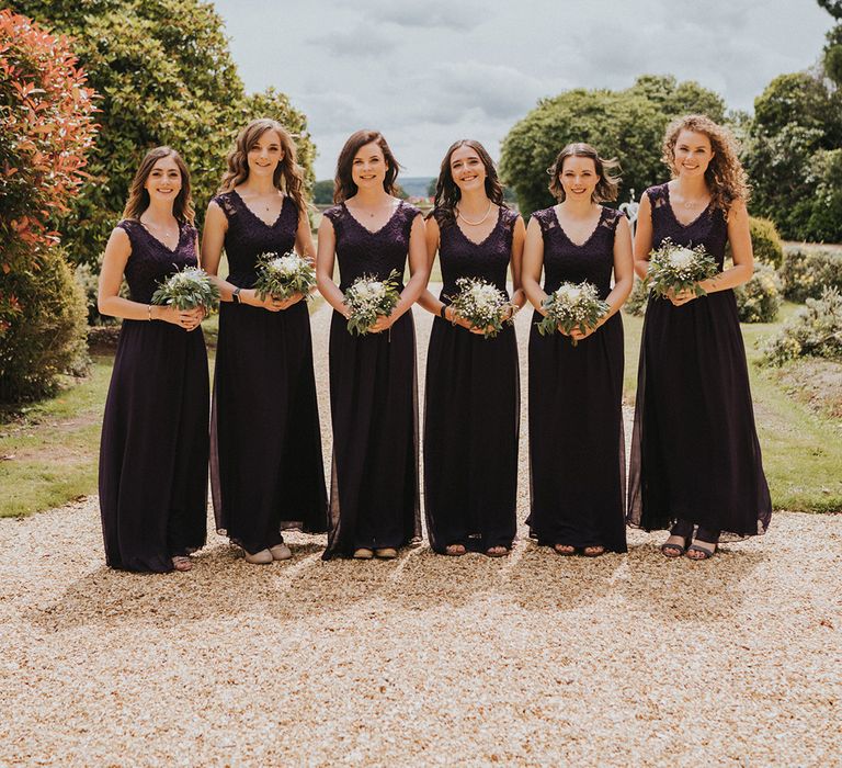 Bridesmaids in deep purple lace dresses holding small bouquet of white roses and gypsophila