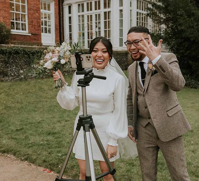 Bride in Vanessa Cocchiaro wedding dress and groom in Tru Clothing outfit posing in front of a phone 