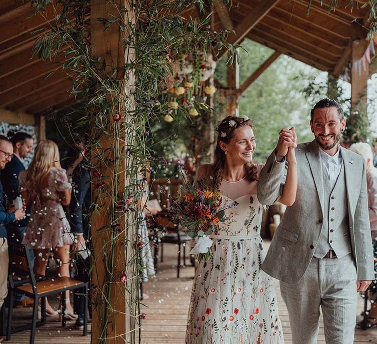 Bride and groom exit their outdoor wedding ceremony with bride wearing floral embroidered dress and groom in grey suit 