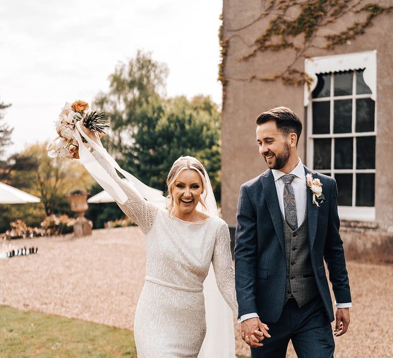 Groom in dark blue suit and paisley tie interlocks hands with bride in beaded dress who lifts her neutral bouquet in the air