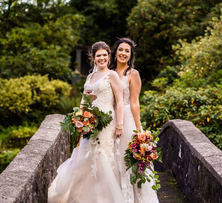 Brides stand together on bridge wearing lace wedding dresses and autumnal wedding bouquets