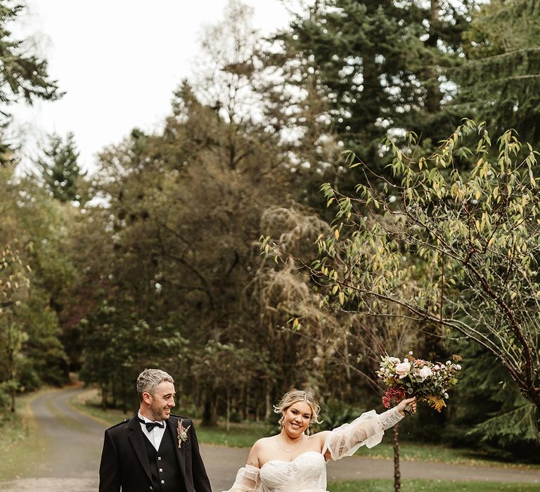 Groom in black tie and green kilt smiles at bride in custom gown lifting her bouquet into the air