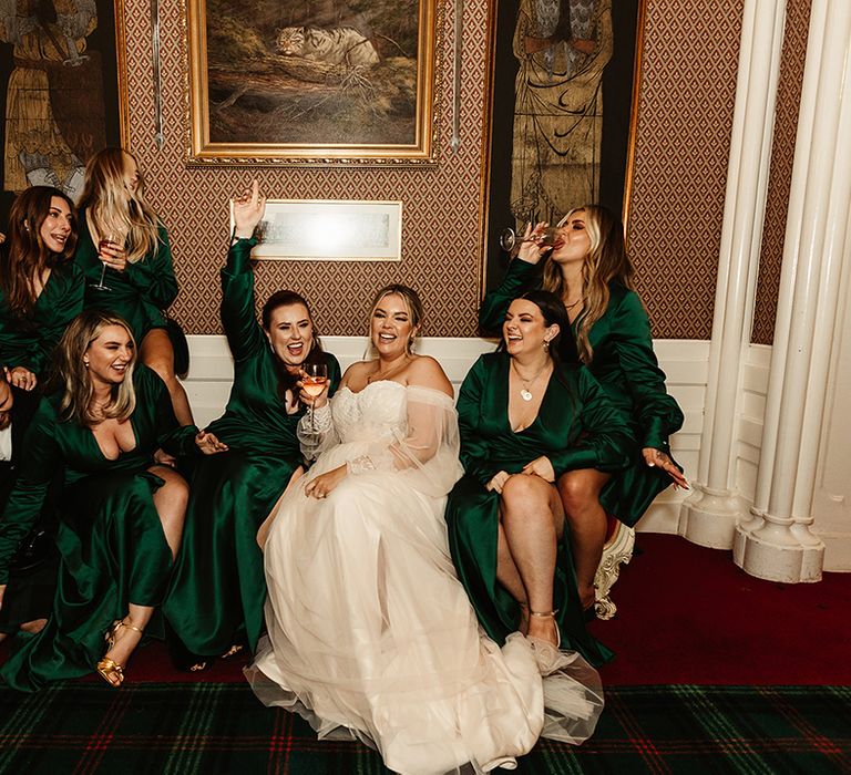 Bride sits holding glass of wine with bridesmaids in emerald green long sleeve dress with front slit also drinking