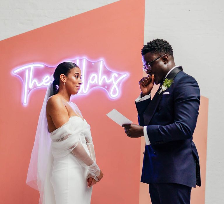 Groom in blue and black tuxedo with shiny black shoes reads out his vows to his bride in front of neon sign and pink background