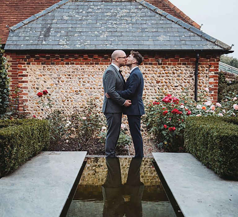 Grooms kiss each other wearing grey and dark blue suits outside their Southend Barns wedding venue 