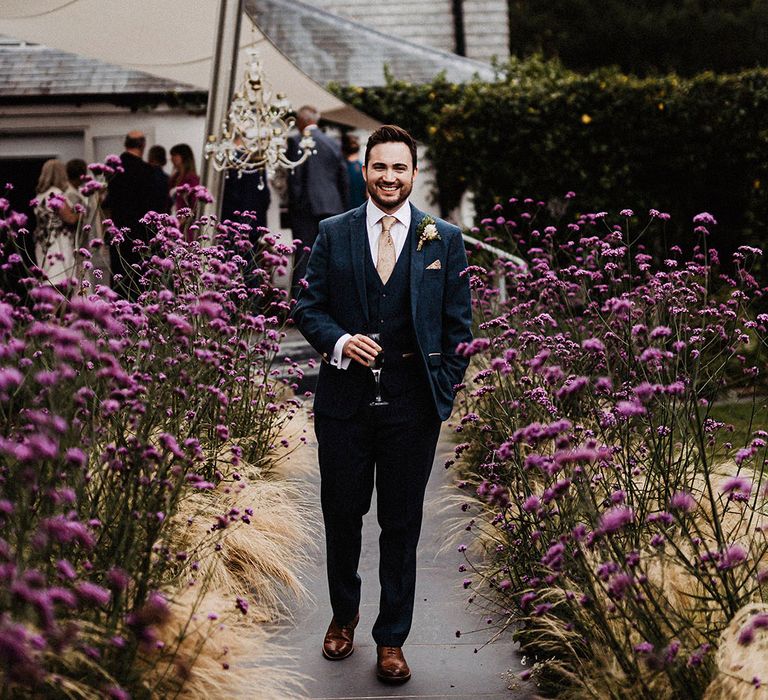 Groom stands next to purple wildflowers wearing blue suit and gold patterned tie and handkerchief