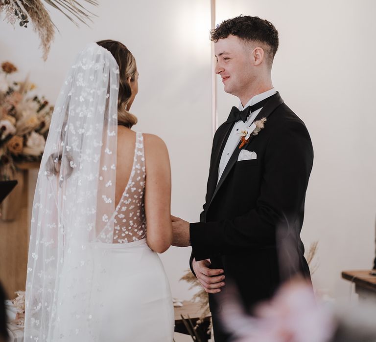 Groom smiles at the altar in black tie and holds hands with bride in Made With Love Bridal gown and matching veil with dot design 