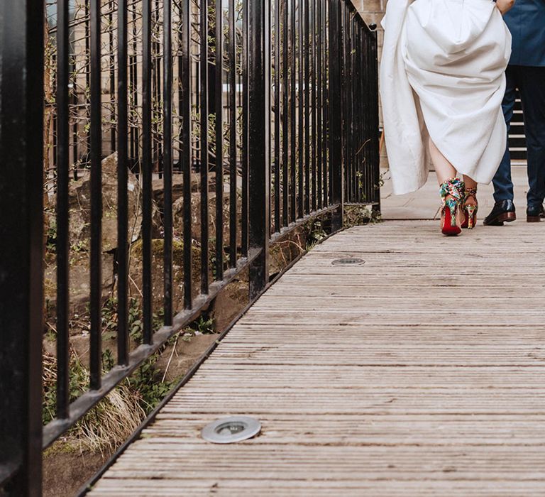 Bride wears bright satin ribbon Louboutin shoes with classic red bottoms walking next to groom in shiny black shoes