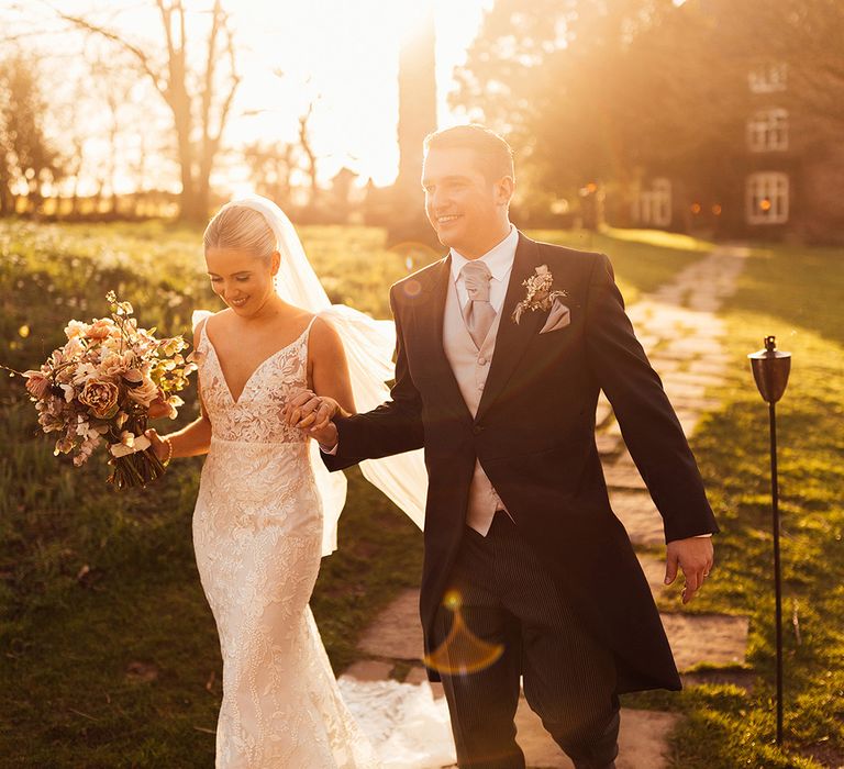 Bride and groom intertwine hands as they walk around Dewsall Court at golden hour 