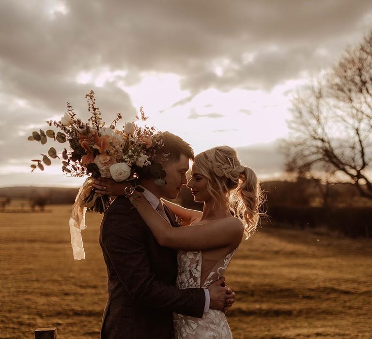 Bride with blonde braided ponytail hairstyle with diamanté clip in Made With Love wedding dress with groom in three piece brown suit