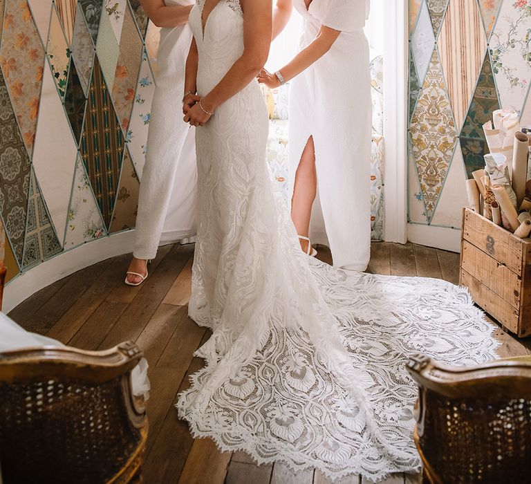 Bridesmaids in sparkly white dresses help the bride into her lace wedding dress with a long train with red lipstick and silver jewellery 