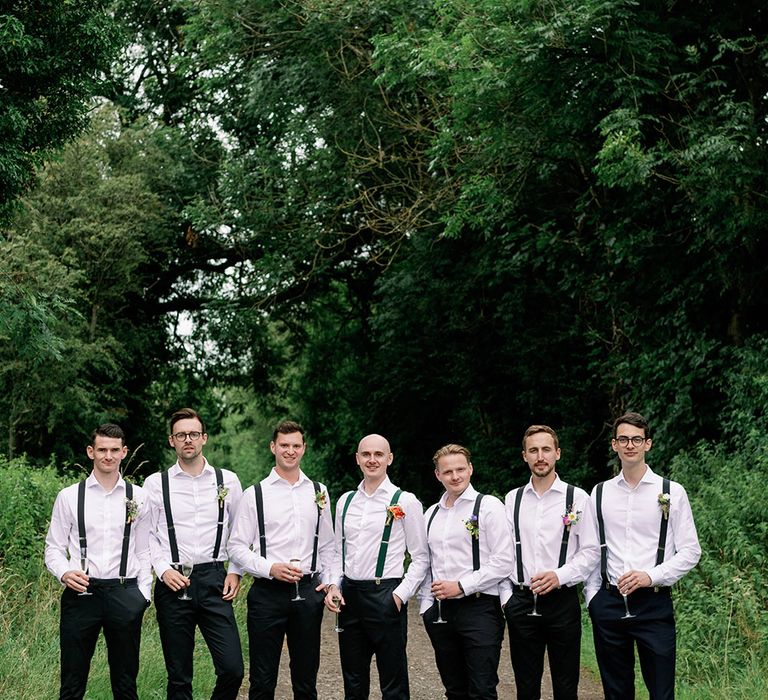 Groom stands with his groomsmen who wear white shirts and green braces 