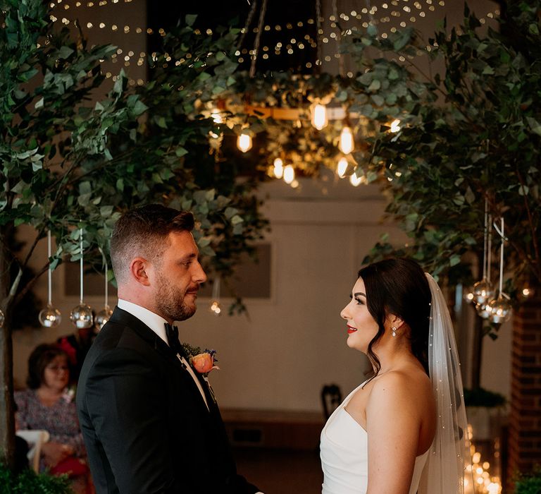 Bride in one shoulder wedding dress with a pearl veil stand with the groom in black tie holding hands at the altar 