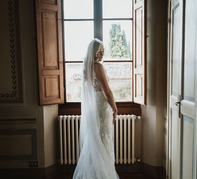 Bride stands in front of large window and wears Emmy Mae Bridal lace wedding dress complete with Made With Love Bridal cathedral veil 