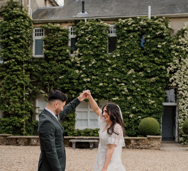 Bride wearing boho floral embellished wedding dress dances with her groom who wears grey suit and green tie 