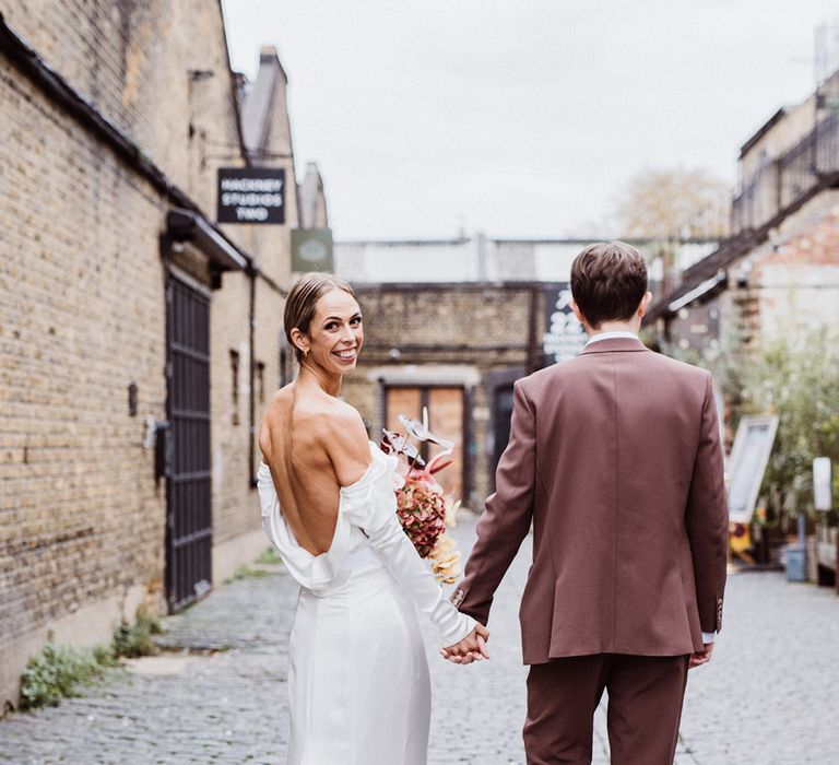 Bride in a cowl neck wedding dress looks back at the camera walking holding hands with the groom in a brown suit