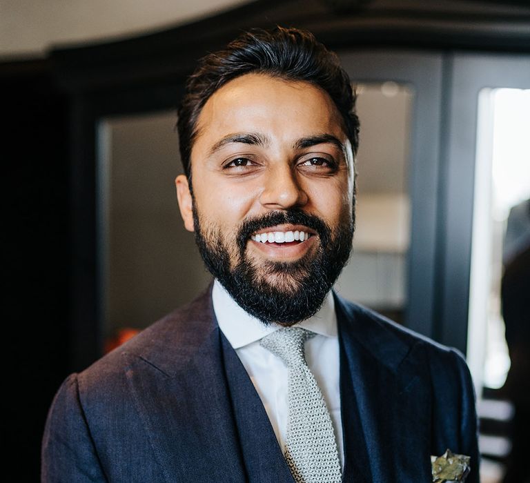 Groom wears three piece blue suit with textured grey tie and floral buttonhole 