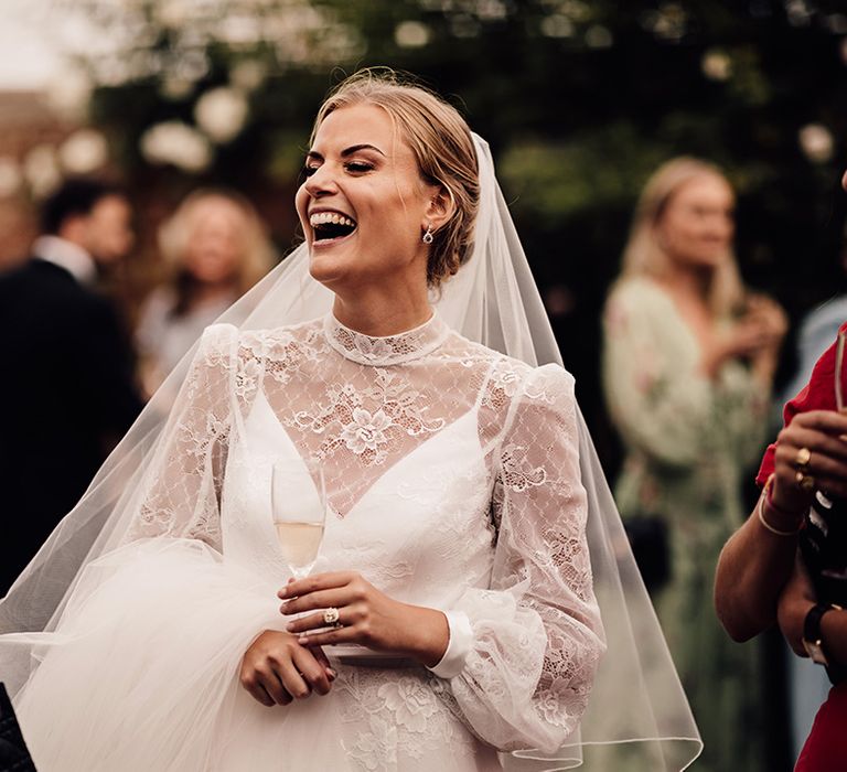 Bride in a long sleeve high neck delicate lace wedding dress holding a glass of champagne laughing with guests 