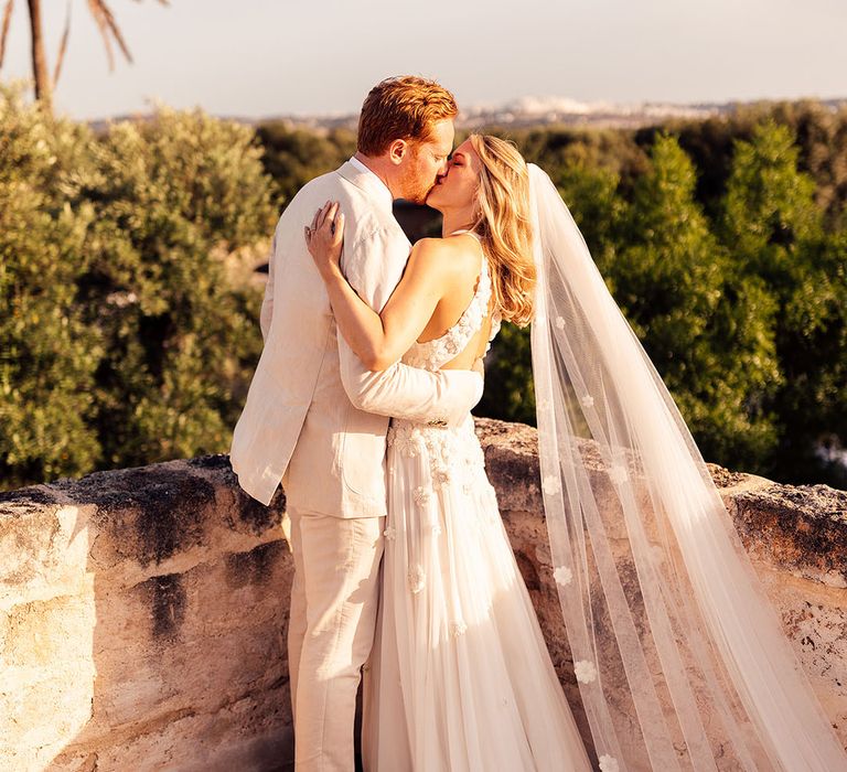 Groom in a cream wedding suit kisses the bride in a flower wedding dress and matching flower veil 