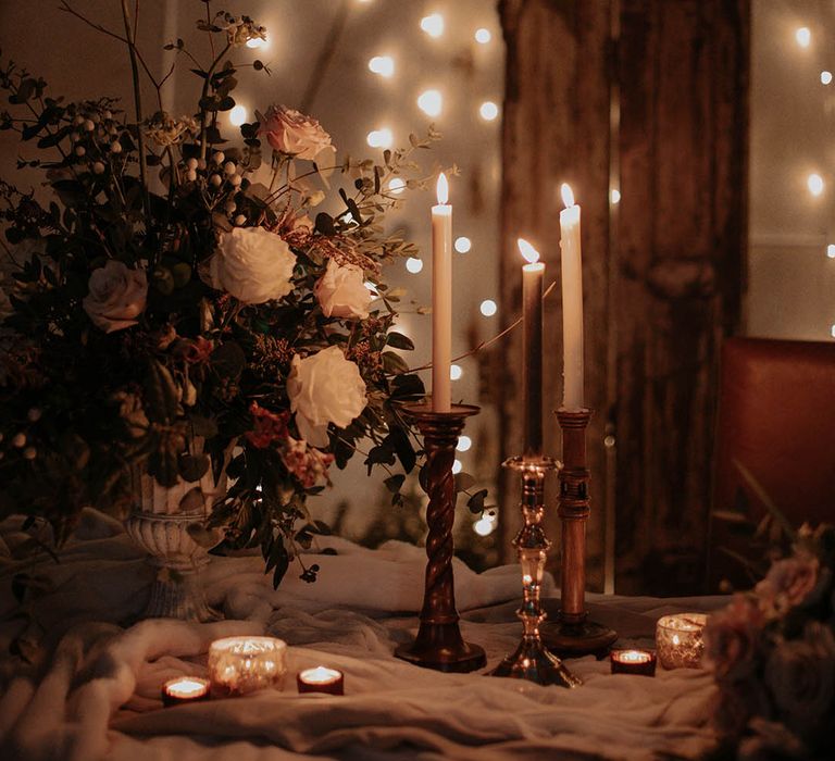 White taper candles in gold candleholders and tea lights with a fairy light backdrop, and white and pink flower arrangement for the table 