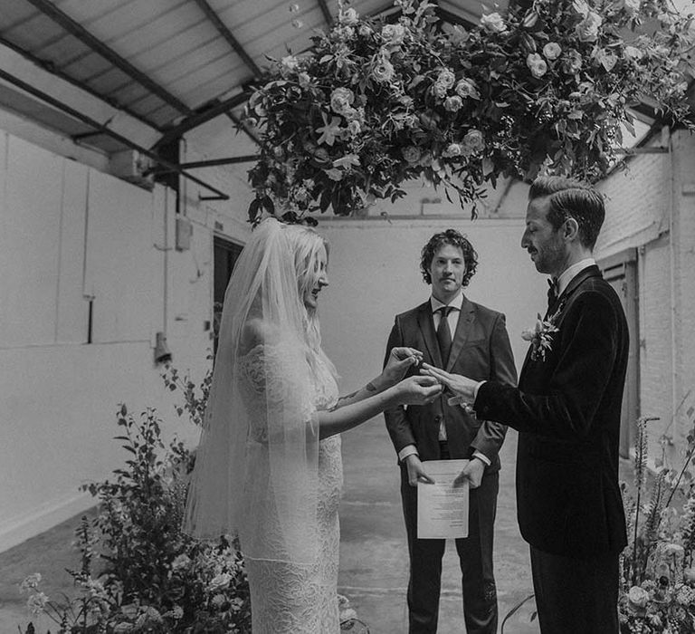 Bride in lace boho two piece with tattoos and groom in black tux with bow tie and boutonniere giving wedding rings at Hackney Studios