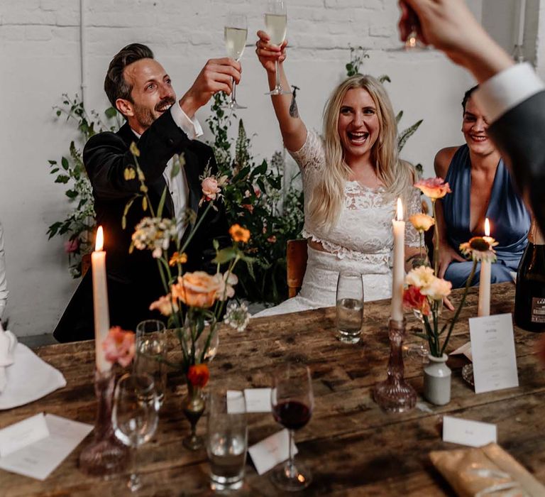 Bride in boho two piece lace bridal outfit and groom in dark tux with bowtie cheering at wedding reception