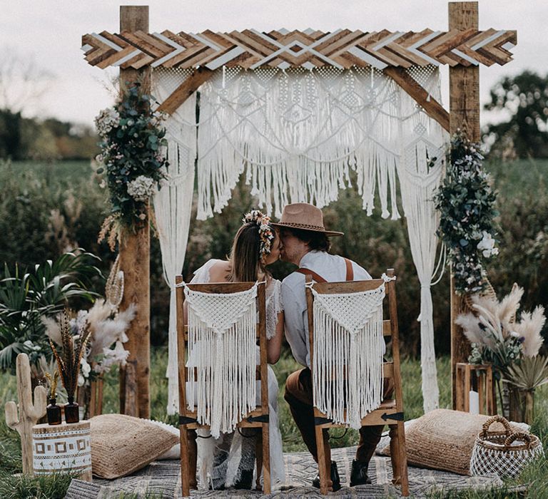 Bride and groom sit on wooden chairs with handmade macrame decor and wooden arch for boho wedding