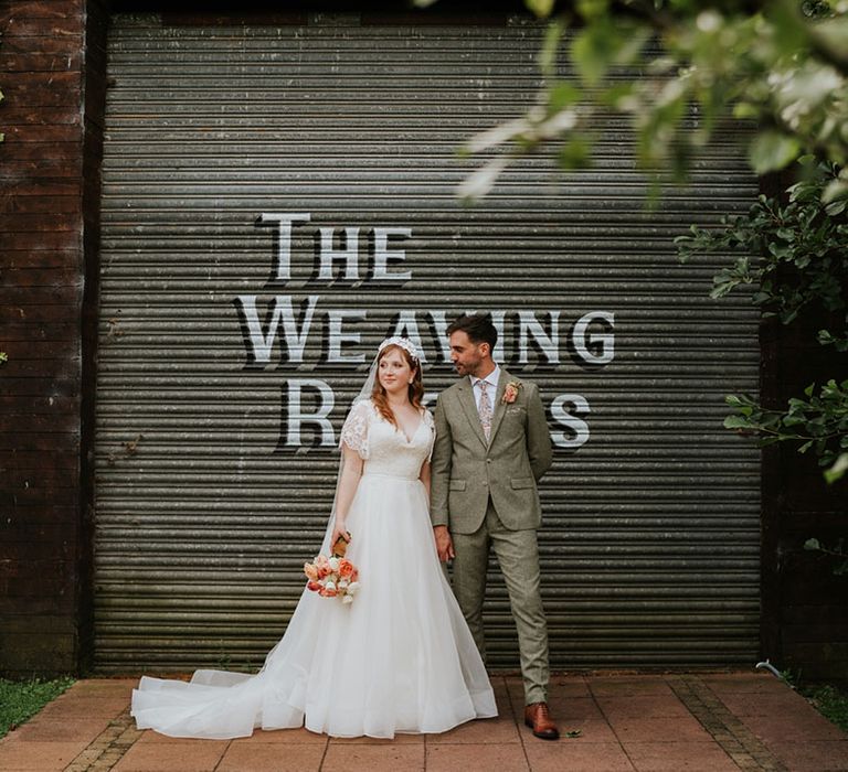 The groom in a tweed suit and floral tie and pocket square smiles at the bride in a lace and tulle wedding dress who holds a peach and coral bouquet